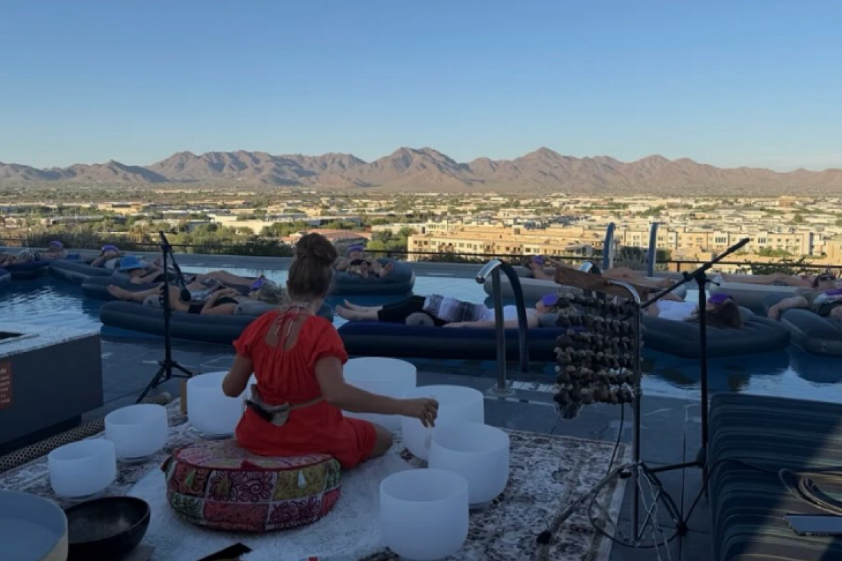 A person in a red dress plays crystal singing bowls for people on pool rafts, a peaceful home preparation scene with mountains in the background.