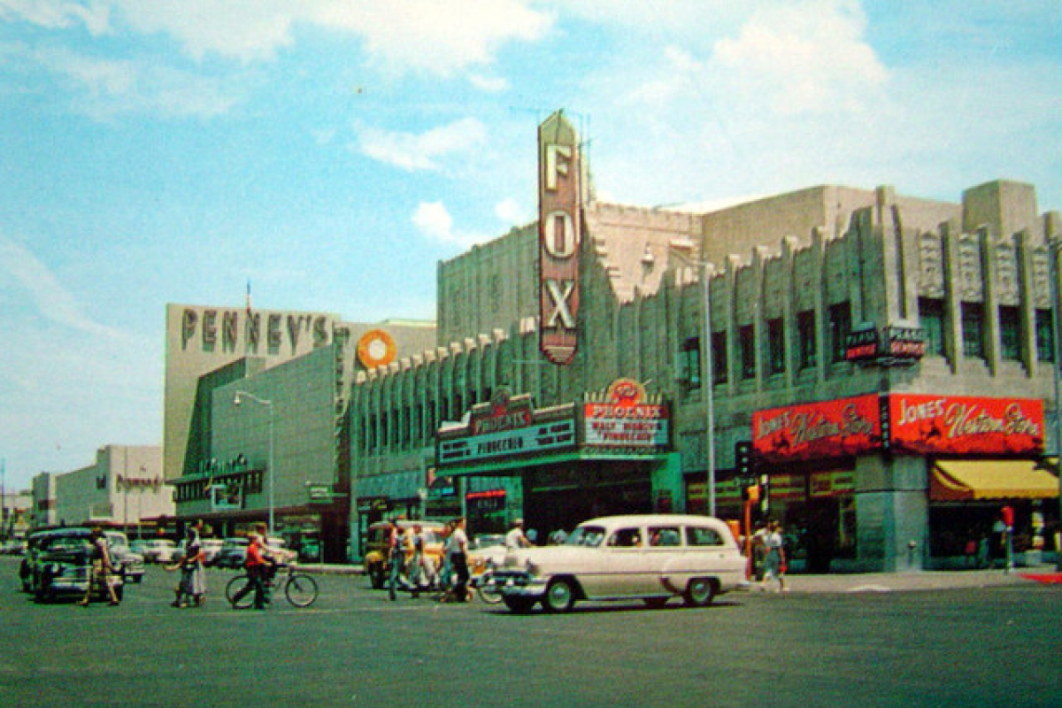 1950s city street with vintage cars, pedestrians, and signs for Phoenix theater, Fox West Coast Theater, and classic stores.