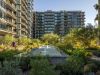 Modern apartment buildings with glass balconies overlook a lush garden and reflecting pool, elevating the Optima living community experience.
