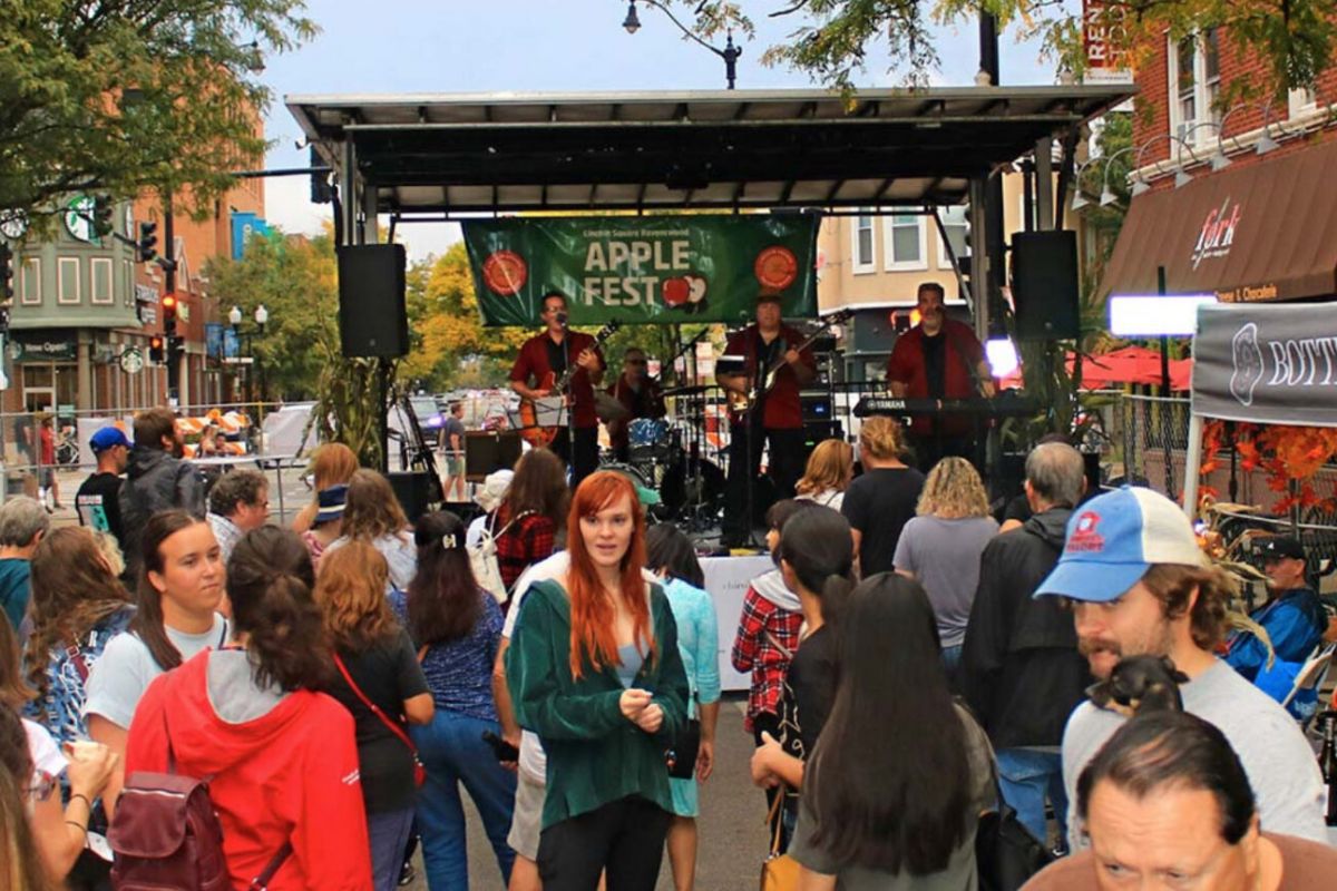 A crowd gathers at an outdoor festival in Chicago with a band playing on stage under a sign that reads "Apple Fest.