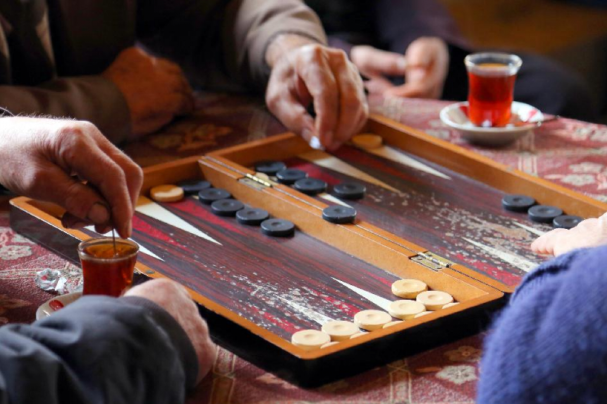 Three people enjoy a fun game of backgammon on a wooden board, with glasses of tea on the table beside them.