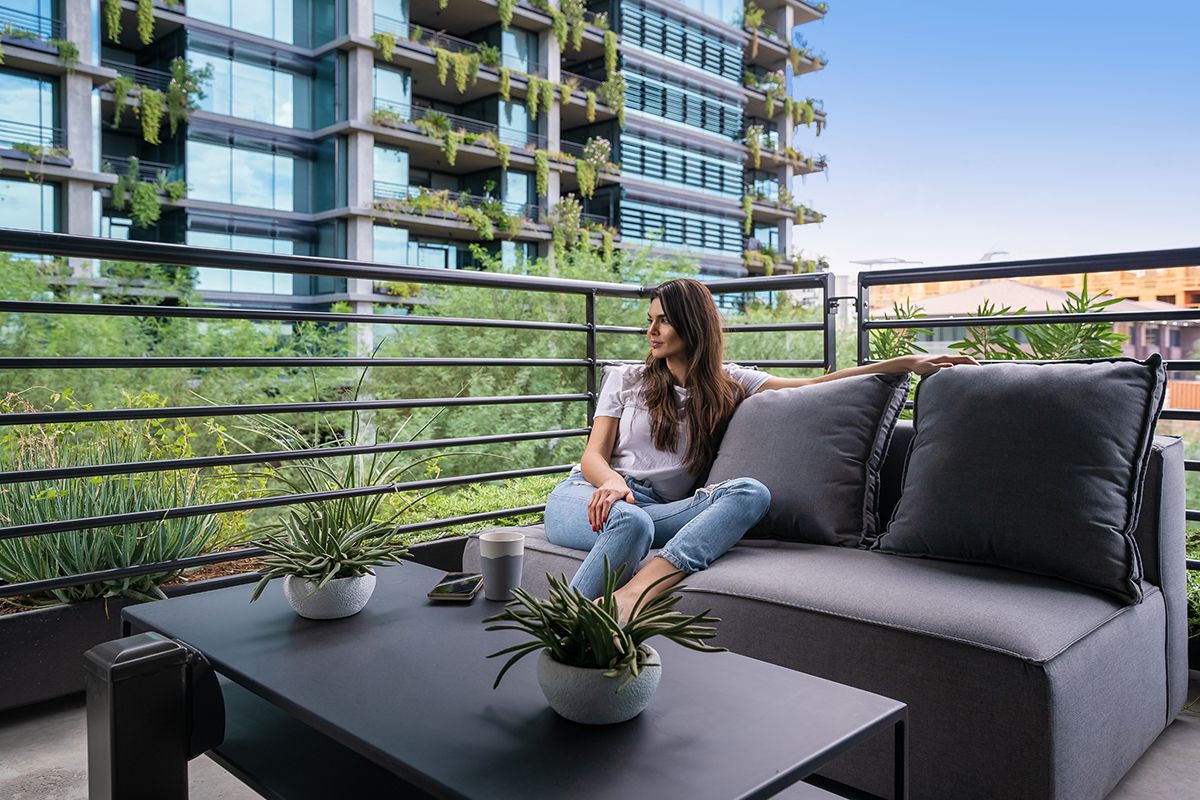 Woman relaxes on a modern balcony sofa with plants, city buildings, and greenery in the background.