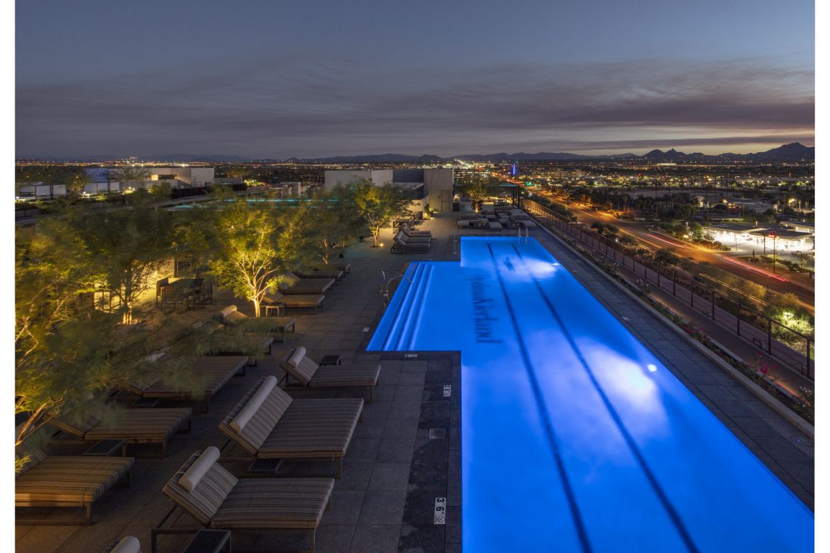 Rooftop pool glowing at dusk with lounge chairs, city lights, and mountains in the background.