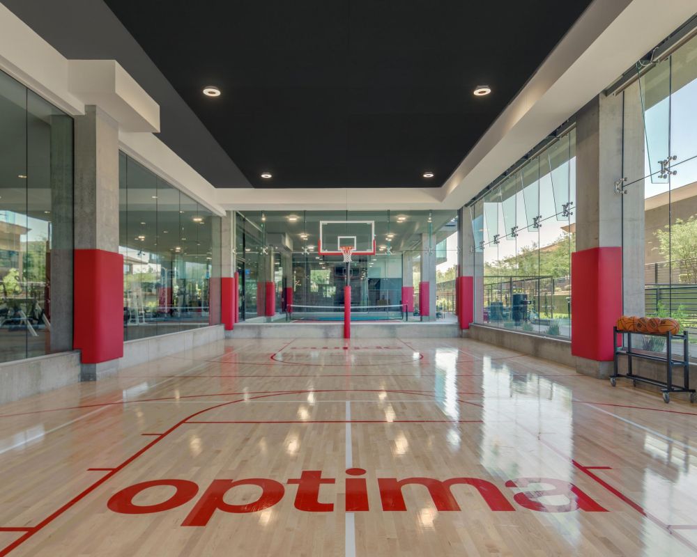 Indoor basketball court with glass walls, red accents, and "optima" logo on the polished wooden floor.