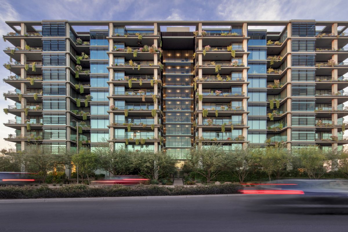 Modern glass apartment with balconies and greenery, showcasing sustainable architecture, viewed from across a street with blurred car lights.