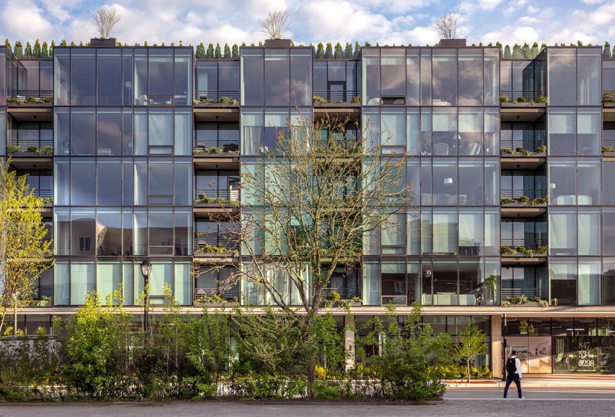 Modern glass building at Optima Verdana apartments with greenery, a tree in front, and a person walking on the sidewalk.