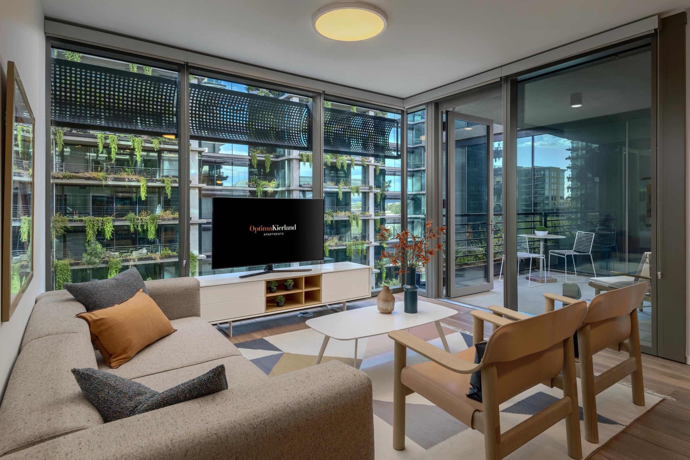 Modern living room with large windows, neutral furniture, a TV, and a view of greenery-covered buildings outside.