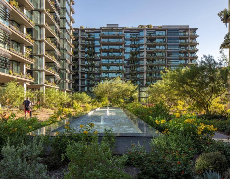 Modern apartment buildings with large windows, garden landscaping, and a central water feature under clear sky.