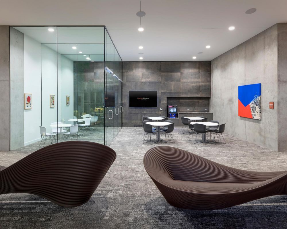 Modern office lounge with sculpted benches, glass meeting room, gray walls, tables, and colorful artwork.