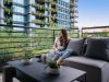 Woman enjoying daily life on a balcony sofa at Optima Community, surrounded by plants, modern buildings, and lush greenery.