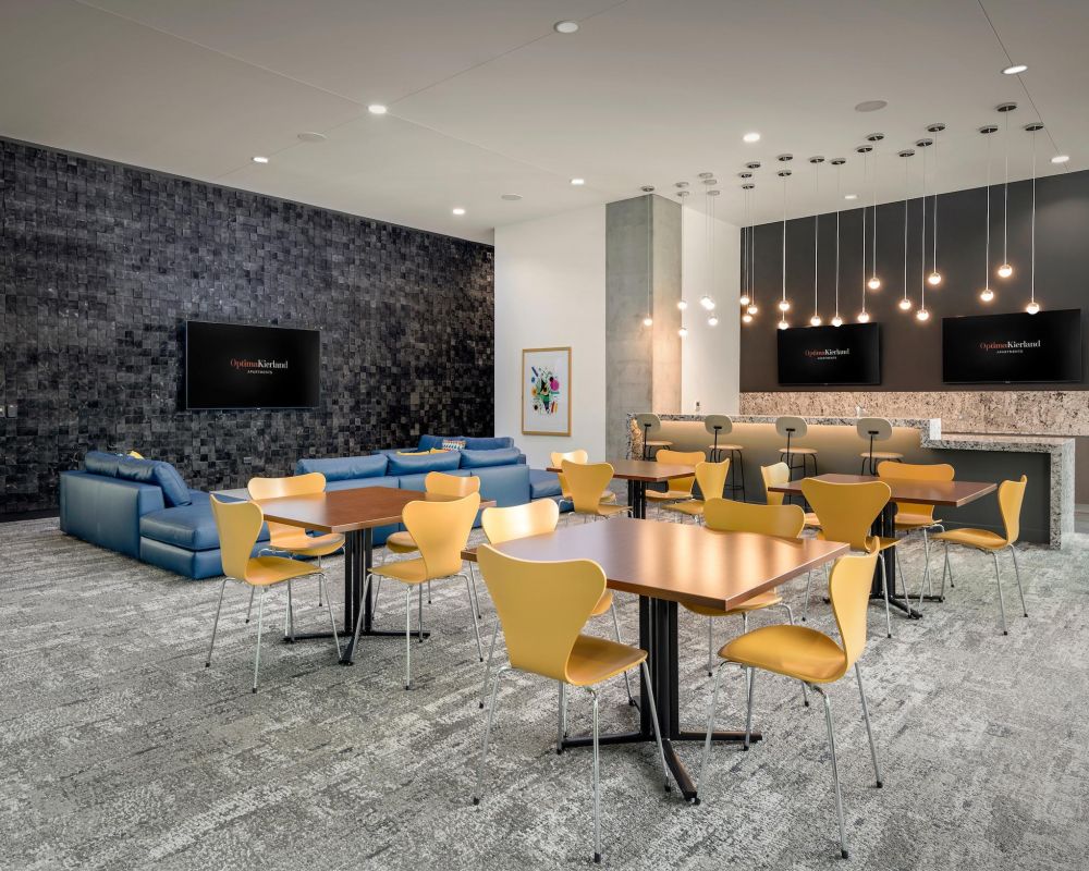 Modern lounge with yellow chairs, wooden tables, blue sofas, and hanging lights, featuring TVs on the walls.
