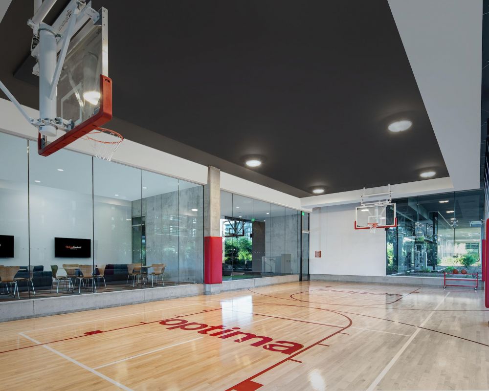 Indoor basketball court with glass walls, red and white markings, and a lounge area with chairs and TVs.