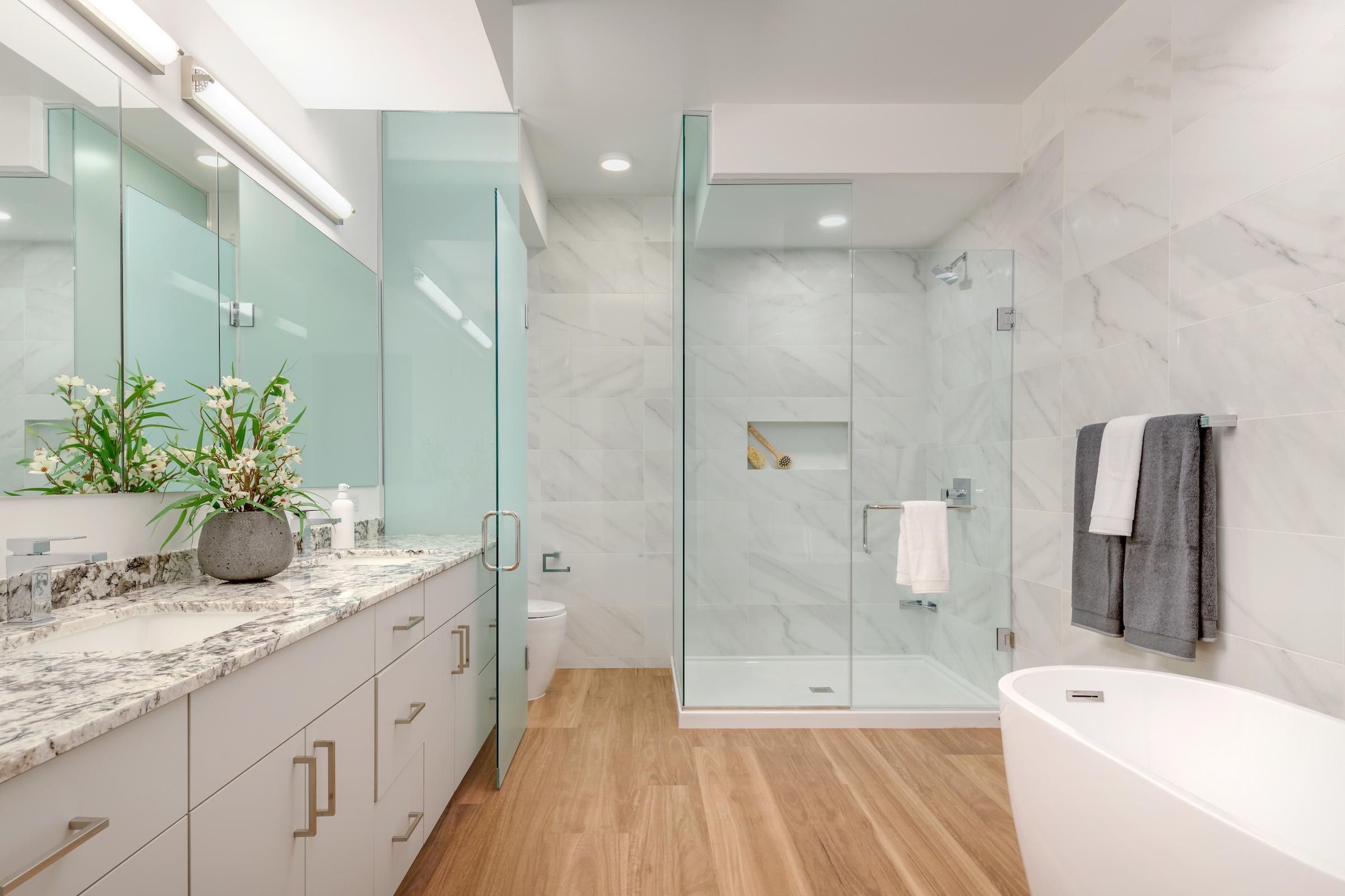 Modern bathroom with marble countertop, glass shower, freestanding tub, and wood flooring.
