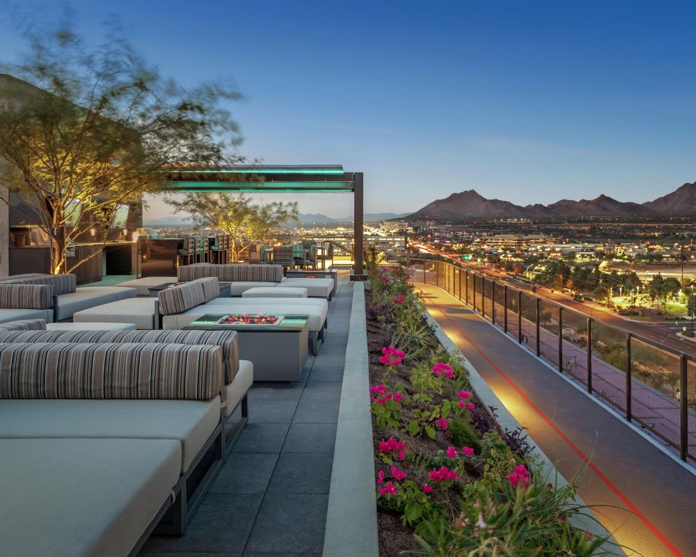 Modern rooftop lounge with striped seating, flower beds, and city and mountain views at sunset.