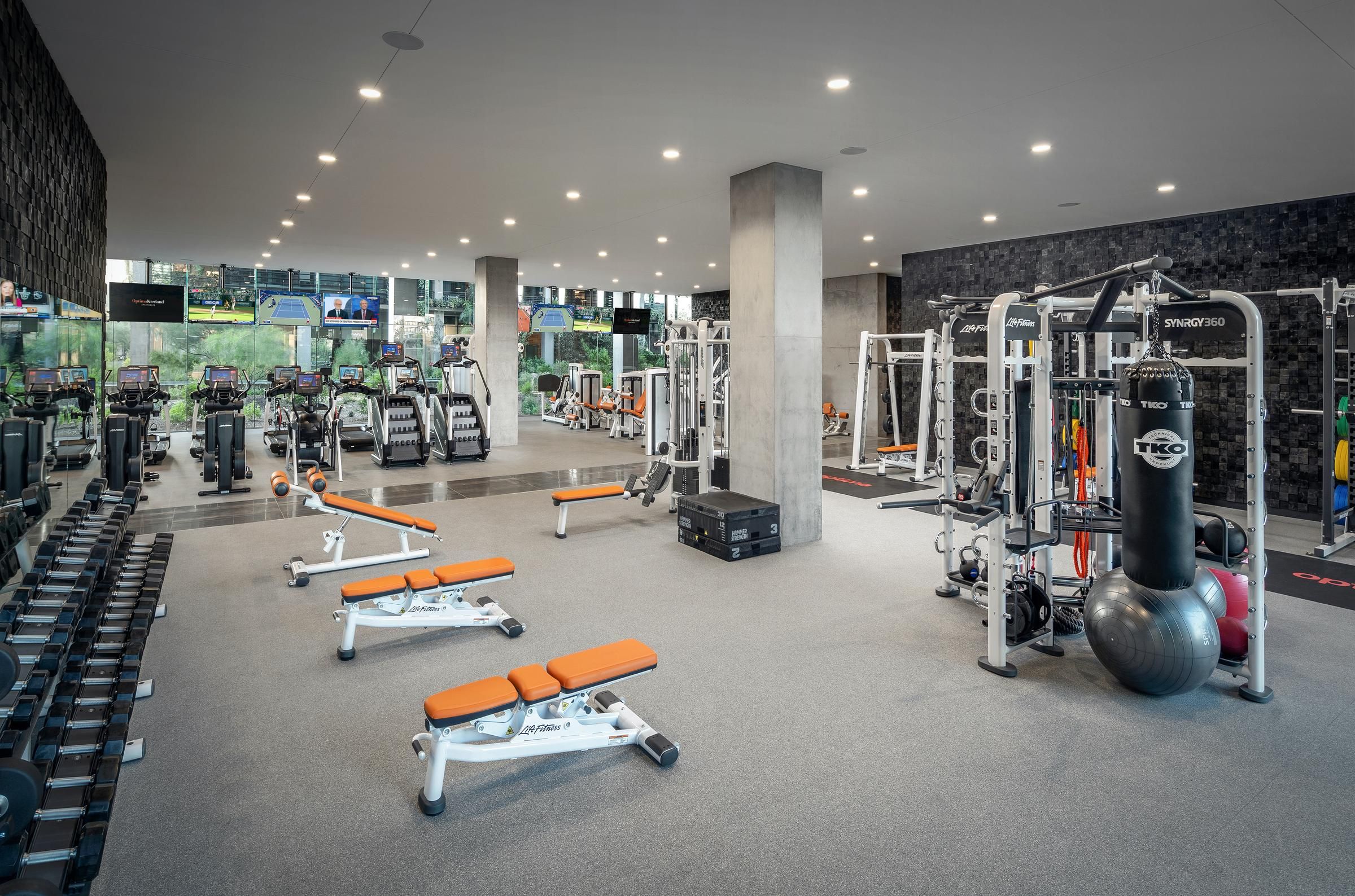 Modern gym with weight machines, free weights, benches, cardio equipment, and large windows with natural light.