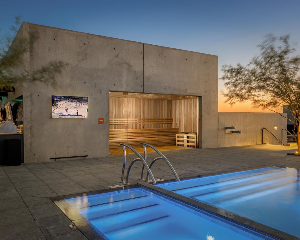 Modern rooftop patio at sunset with a lit pool, a TV showing basketball, and a sauna in a concrete building.