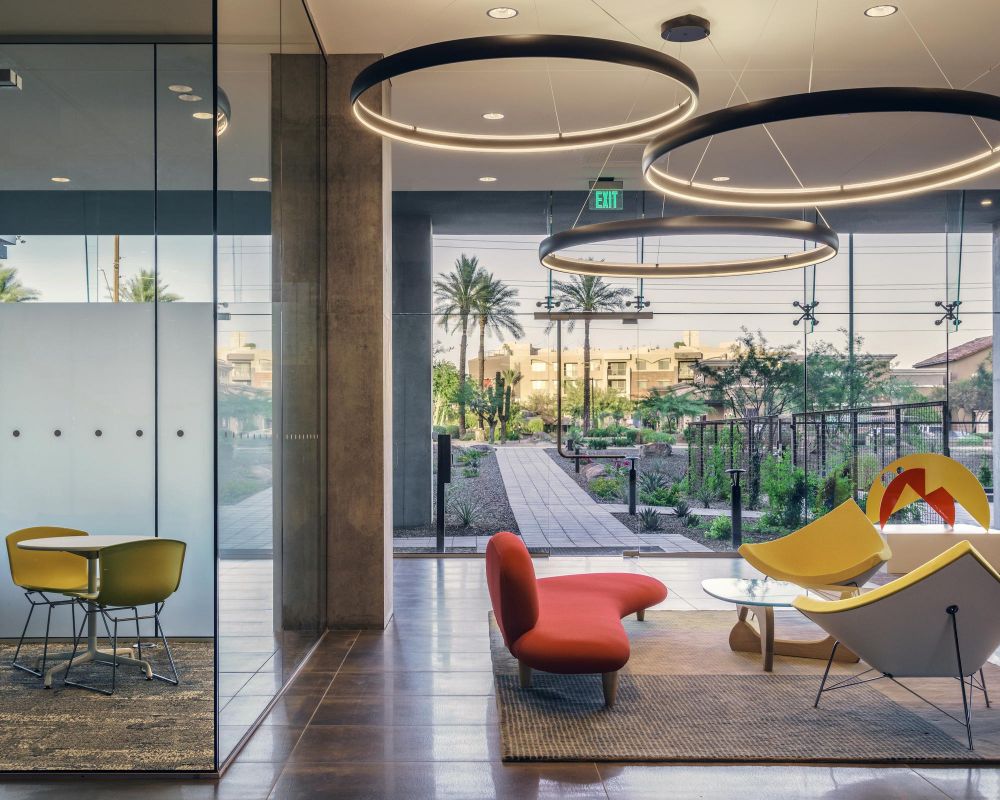 Modern office lounge with colorful chairs, large circular lights, and a glass wall overlooking a landscaped courtyard.
