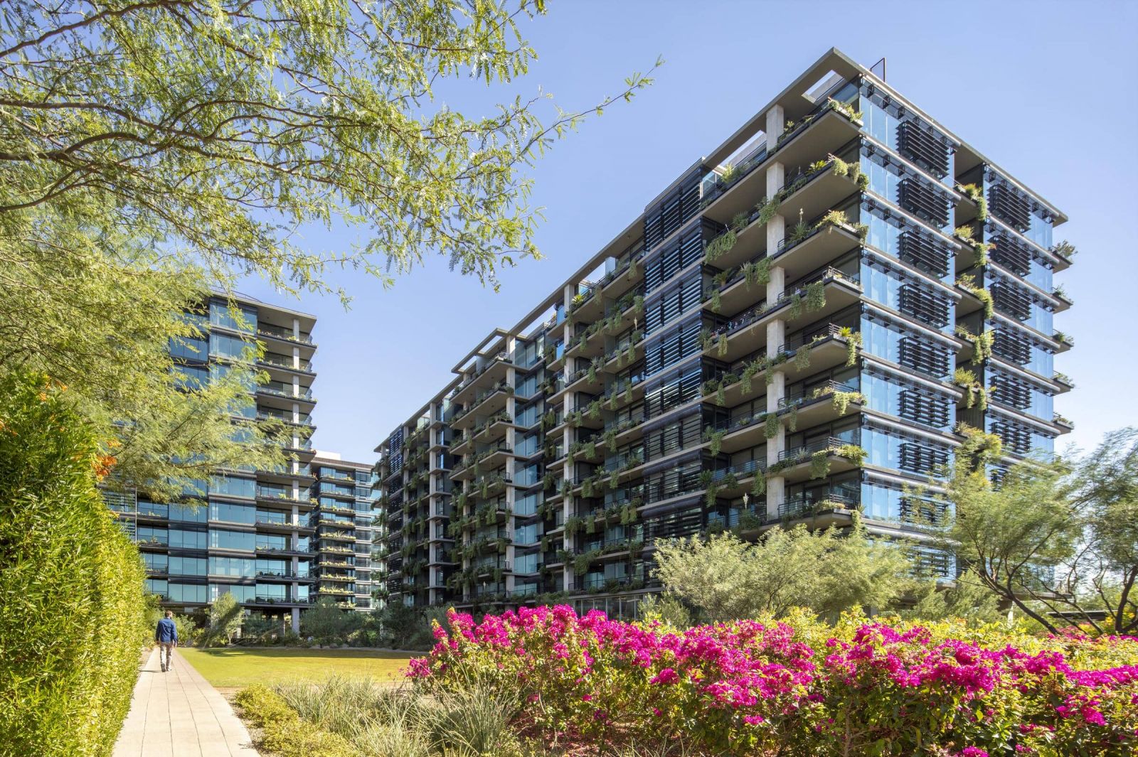 Modern apartment buildings with balconies, surrounded by greenery and colorful flowers under a clear blue sky.