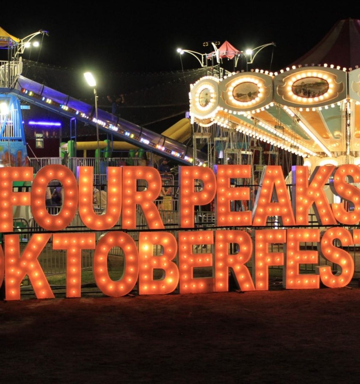 Brightly lit "Four Peaks Oktoberfest" sign at Tempe Beach Park with carnival rides glowing at night in the background.