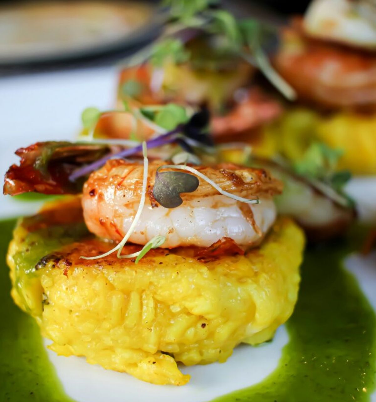 A shrimp sits atop a yellow rice cake, garnished with microgreens and green sauce—Savor the Flavor at Arizona Restaurant Week, Spring 2024.