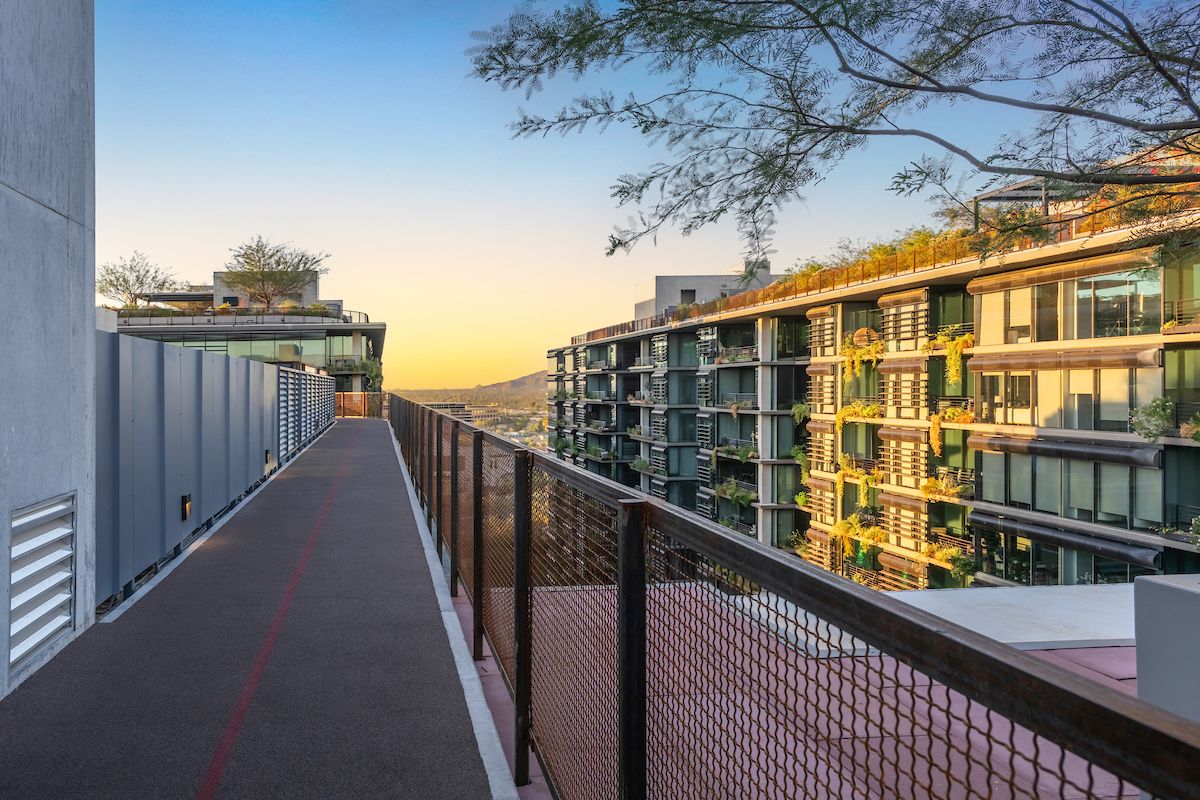 Sunlit walkway with railings between modern apartment buildings, ideal for morning rituals or daily routines amid trees and a distant mountain view.