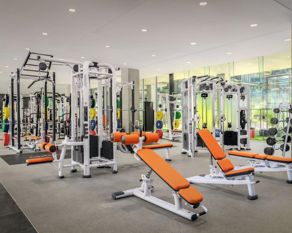 Modern gym with orange benches, weight machines, dumbbells, and large mirrors reflecting the bright space.