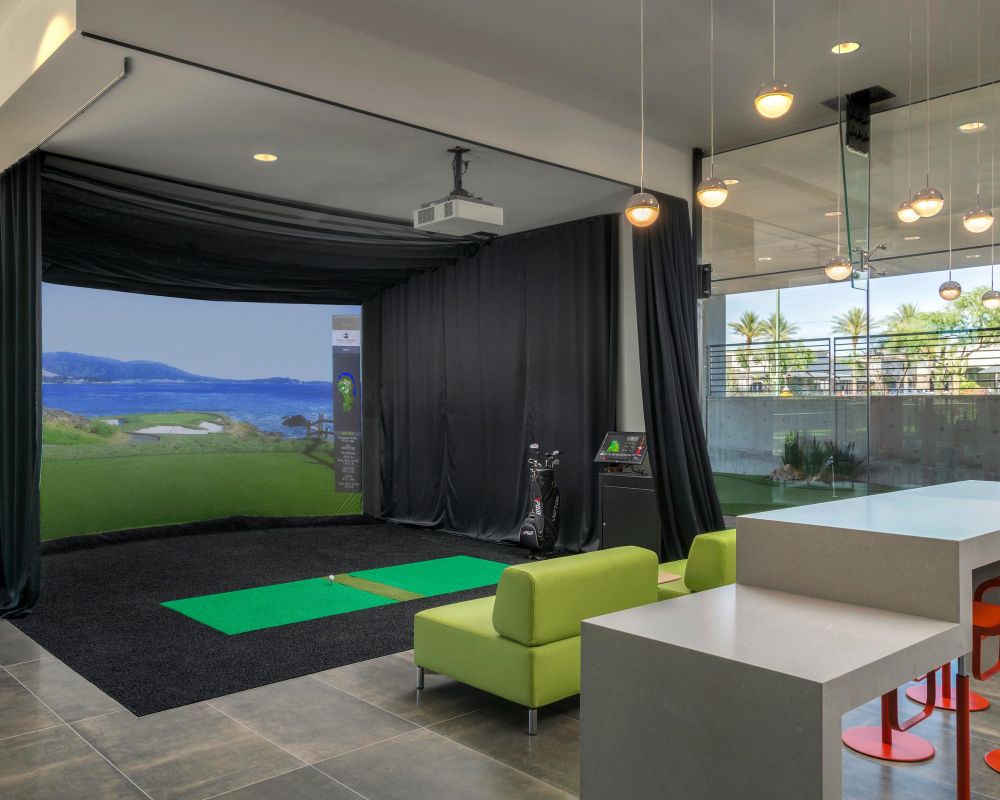 Modern indoor golf simulator setup with a green mat, lounge seating, and bar stools in a stylish room.