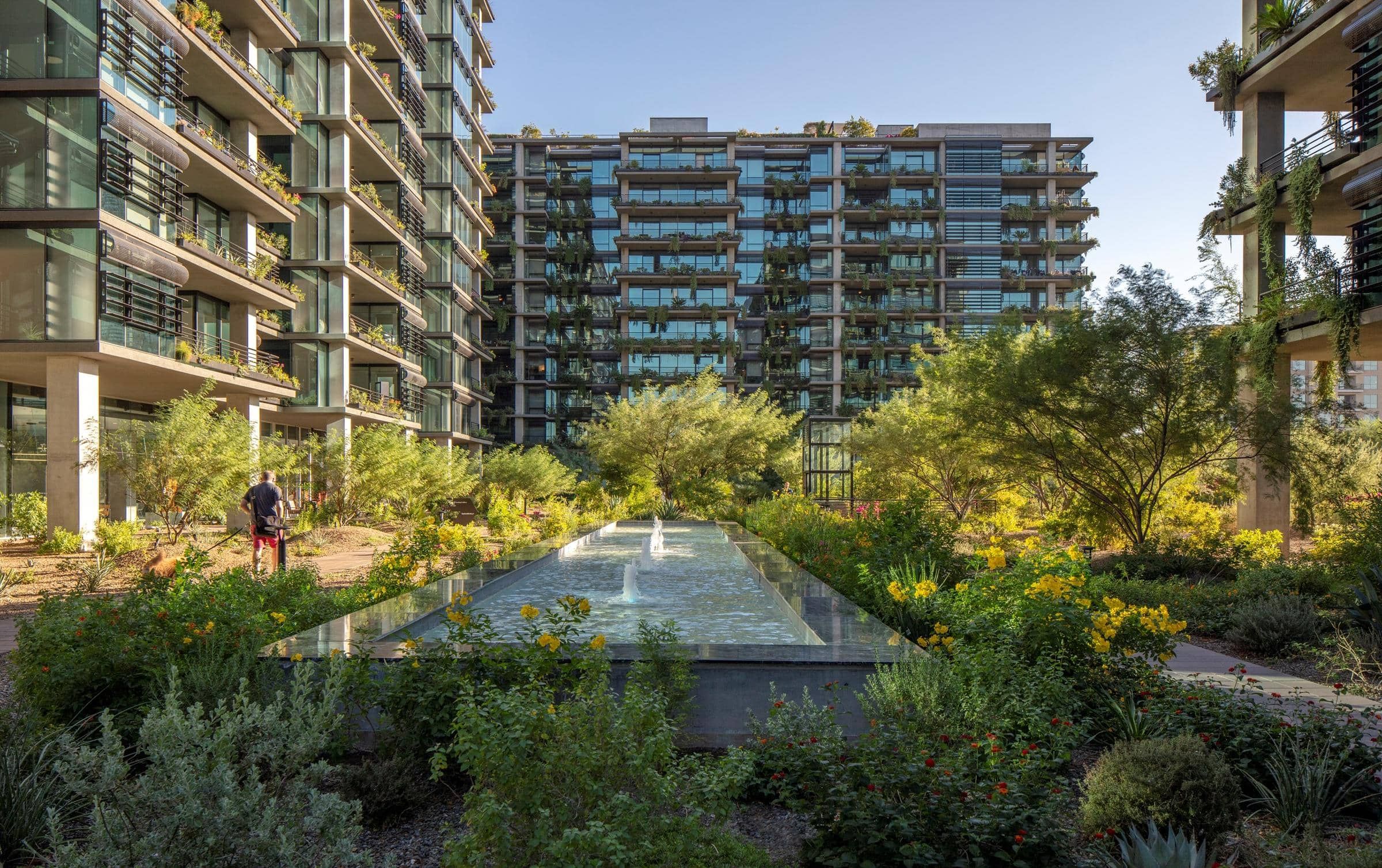 Modern apartment buildings with large windows, garden landscaping, and a central water feature under clear sky.