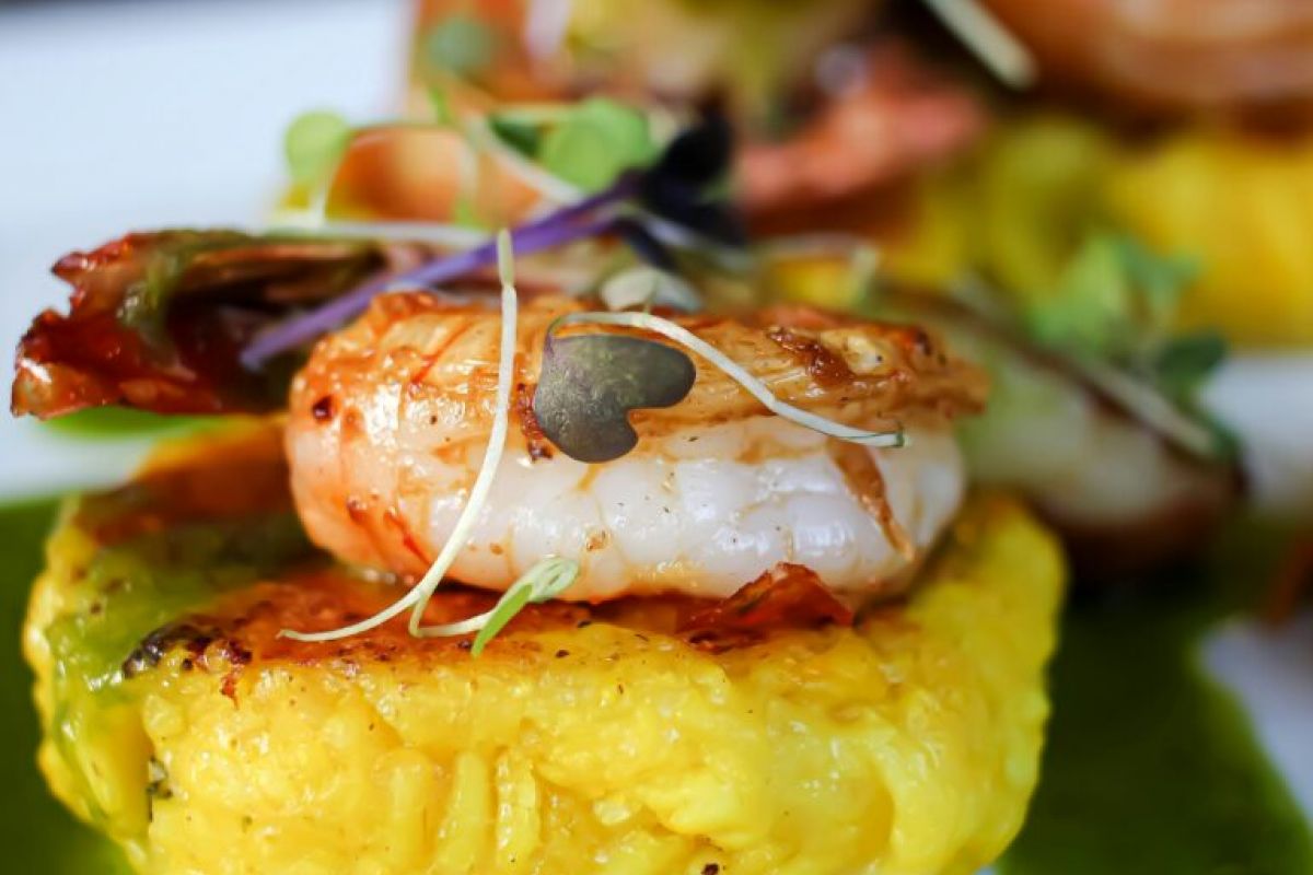 A shrimp sits atop a yellow rice cake, garnished with microgreens and green sauce—Savor the Flavor at Arizona Restaurant Week, Spring 2024.