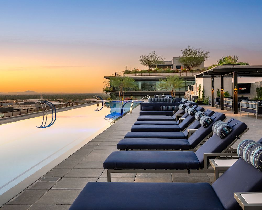 Rooftop infinity pool with lounge chairs at sunset, overlooking a city and distant mountains.