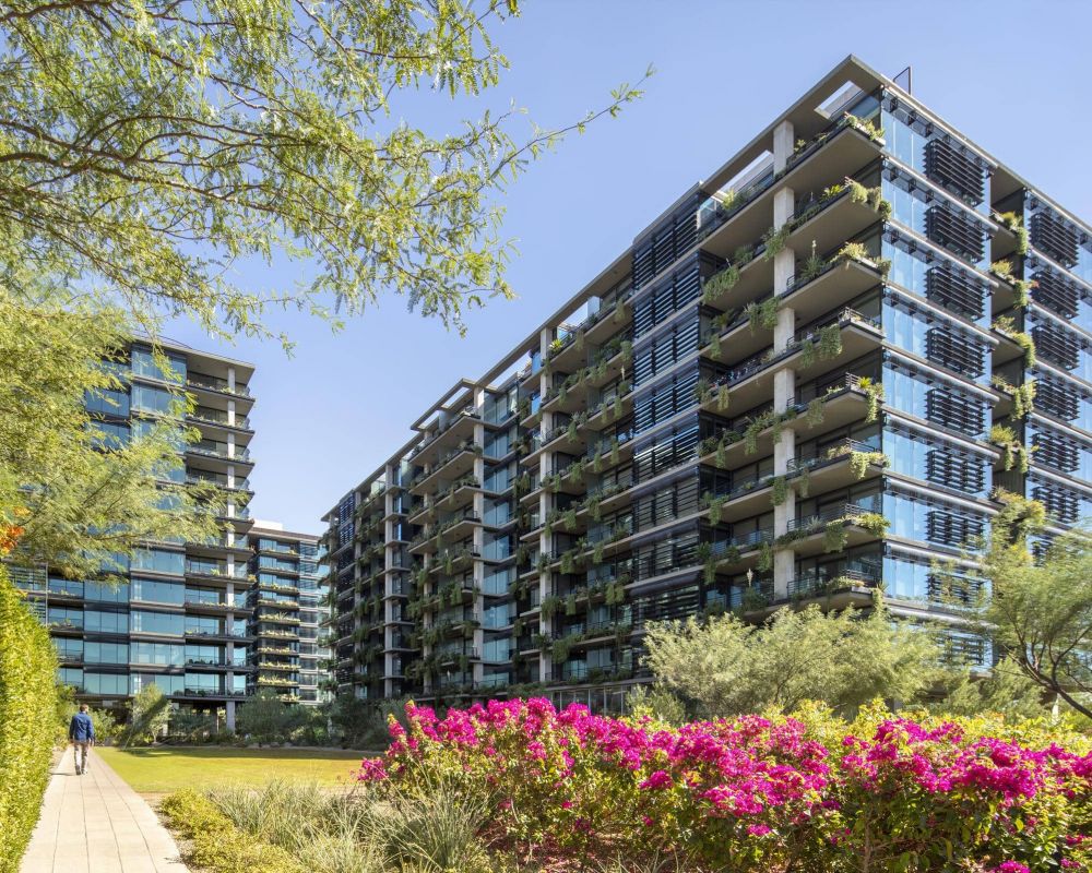 Modern apartment buildings with balconies, surrounded by greenery and colorful flowers under a clear blue sky.