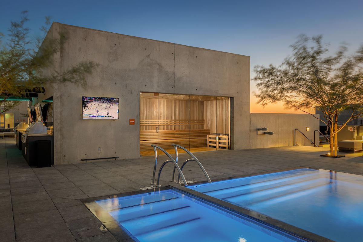 Modern rooftop at Optima Kierland Apartments with a glowing pool, sauna for health benefits, TV showing sports, and trees at sunset.