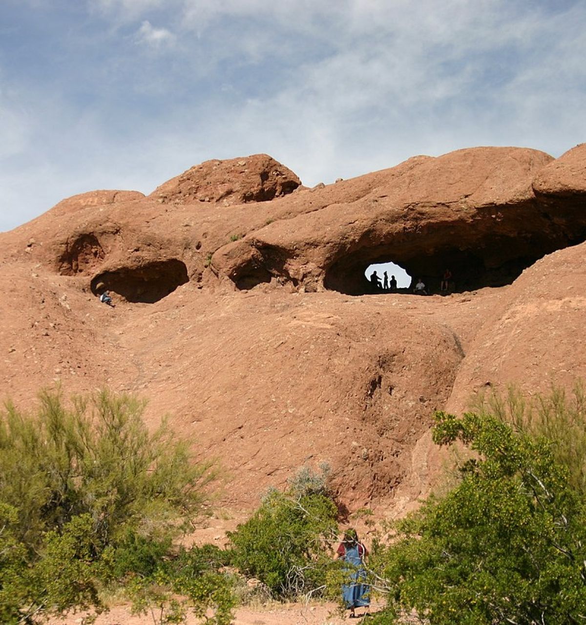 Reddish rock formation with large openings, people inside, and desert vegetation—perfect for hiking along Papago Park trails.
