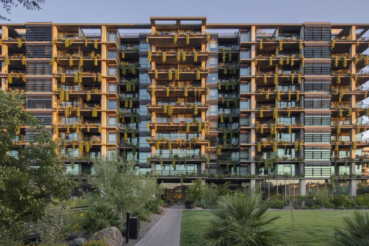 Modern apartment building with many balconies decorated with hanging plants, surrounded by greenery.