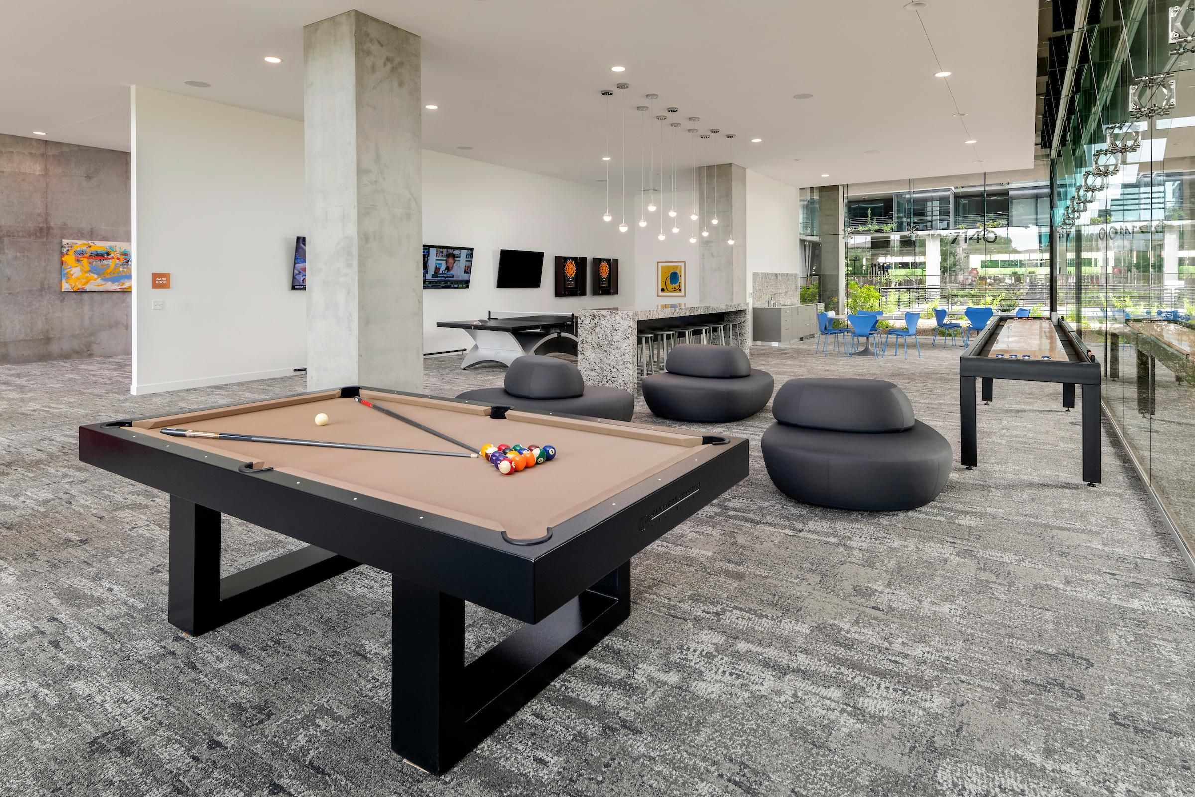 Modern game room with a pool table, lounge chairs, wall art, and large windows overlooking a courtyard.