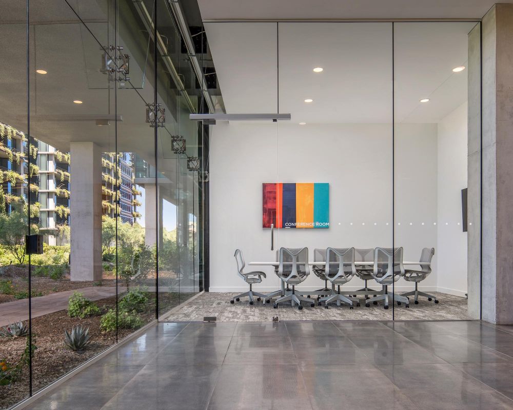 Modern glass-walled conference room with gray chairs, colorful wall art, and a garden view outside.