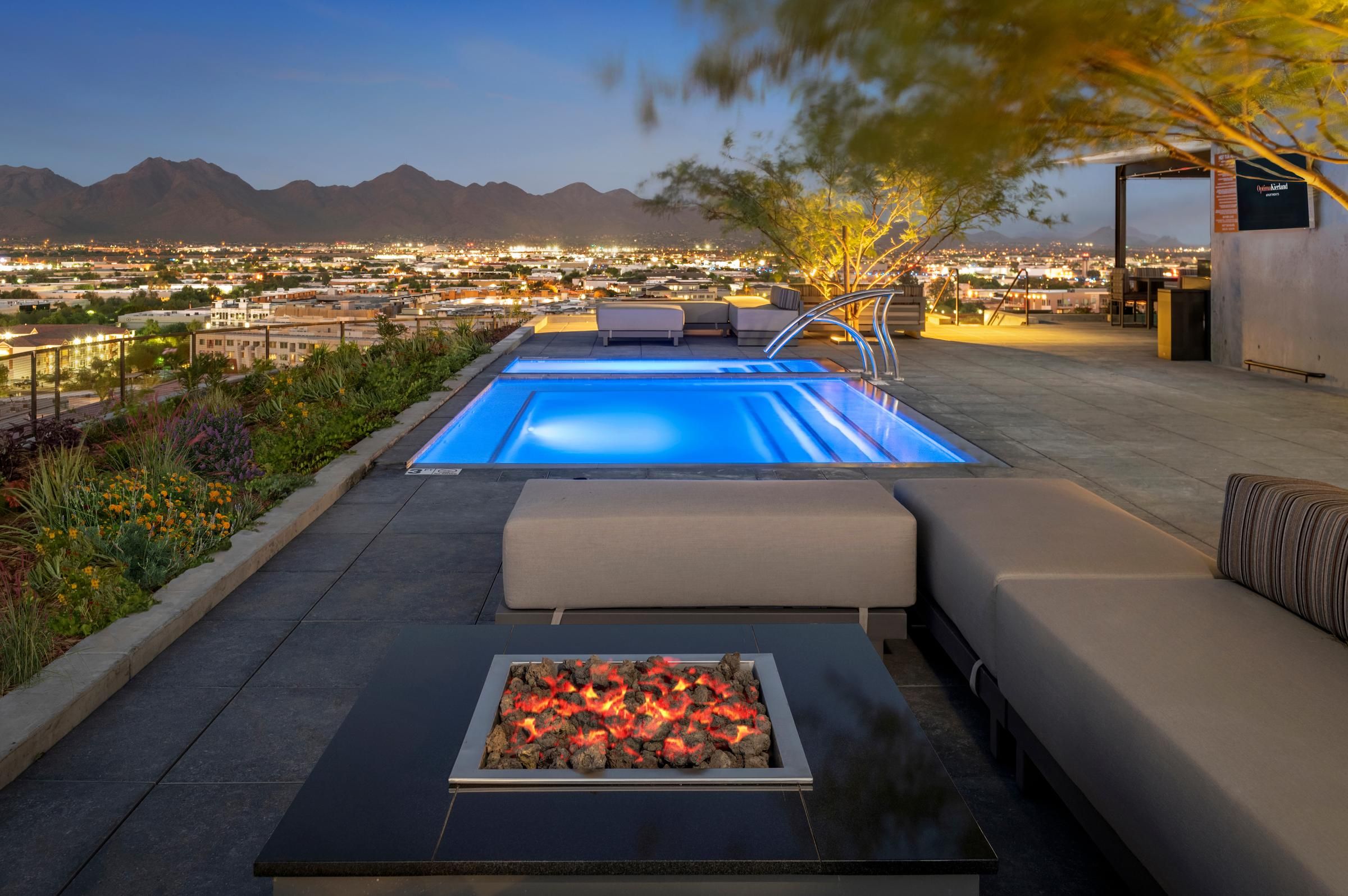 Modern rooftop patio with a fire pit, pool, and city view at sunset, mountains visible in the background.