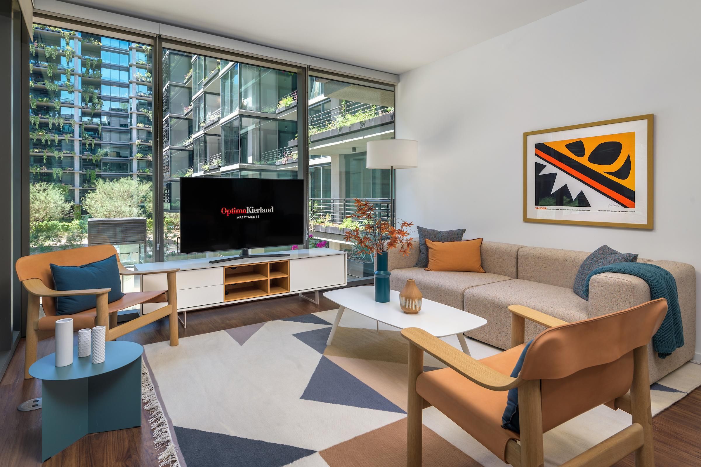 Modern living room with large windows, neutral furniture, abstract art, and view of green apartment buildings outside.