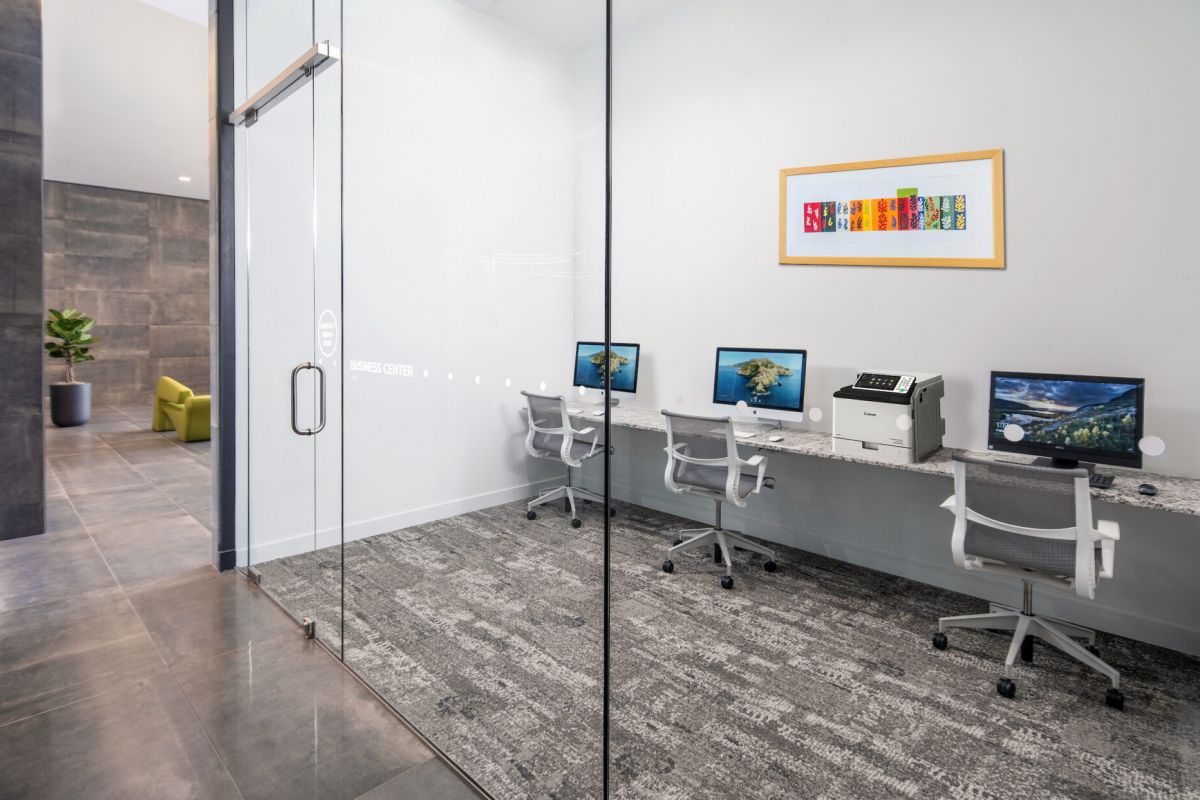 A modern business center by Optima Communities with three computers, a printer, and office chairs—perfect for remote working behind a glass door.