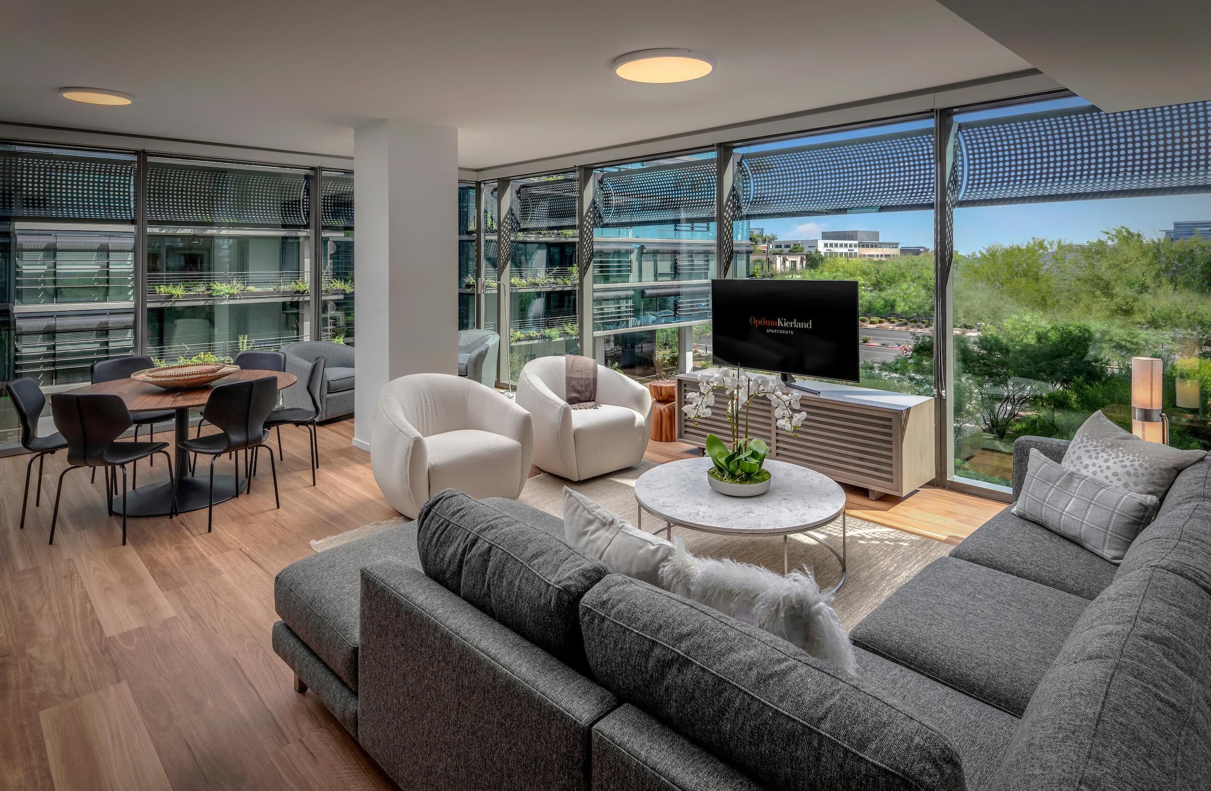 Modern living room with large windows, gray sofa, armchairs, dining area, and cityscape view outside.