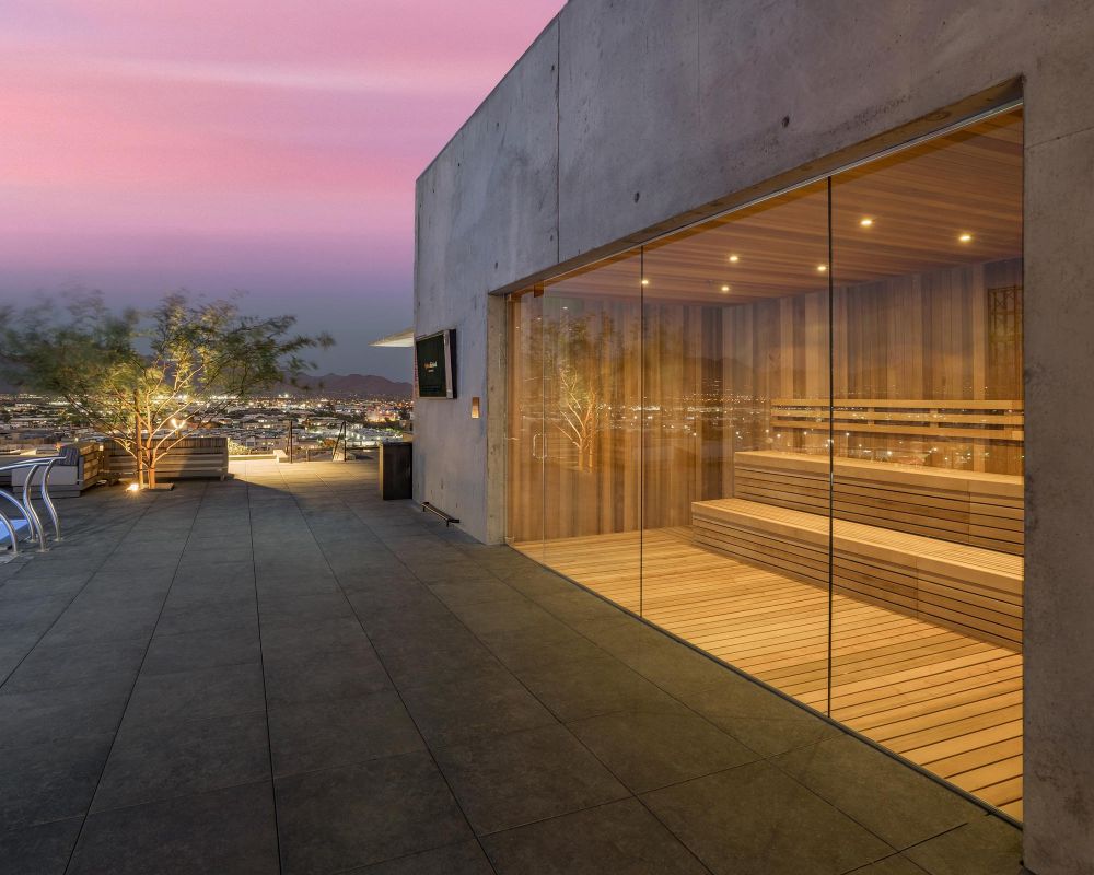 Rooftop sauna with glass walls overlooking a cityscape at sunset, near a pool and lounge chairs.