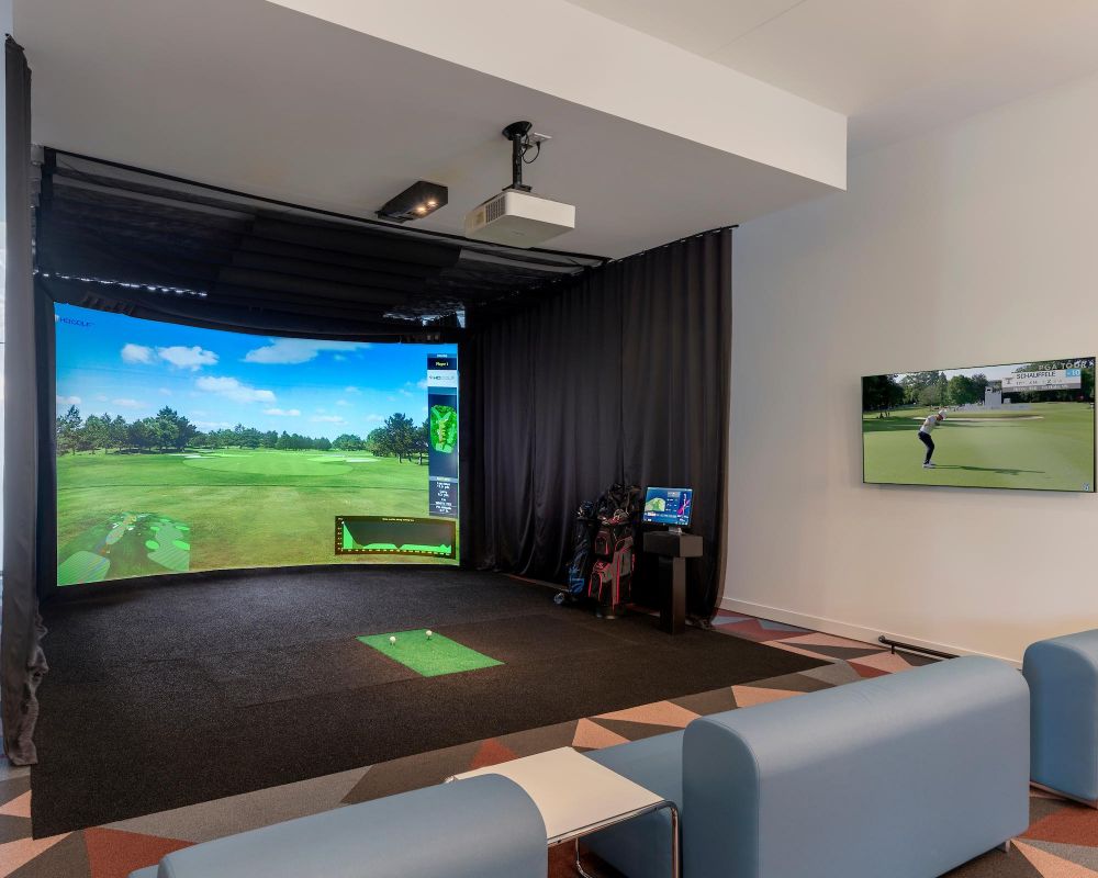 Indoor golf simulator setup with a large screen, golf clubs, and lounge seating in a modern room.