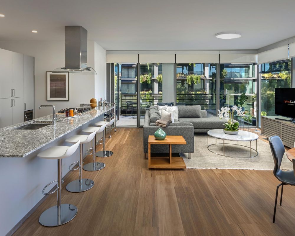 Modern open-plan kitchen and living area with bar stools, gray sofa, and large windows letting in natural light.