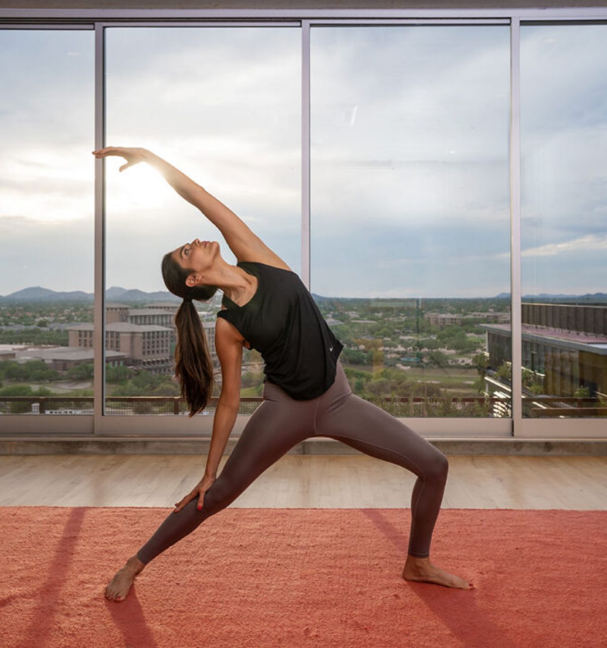 Woman practicing yoga in a modern room, embracing mindfulness as she stretches on a red mat before large windows with a city view.
