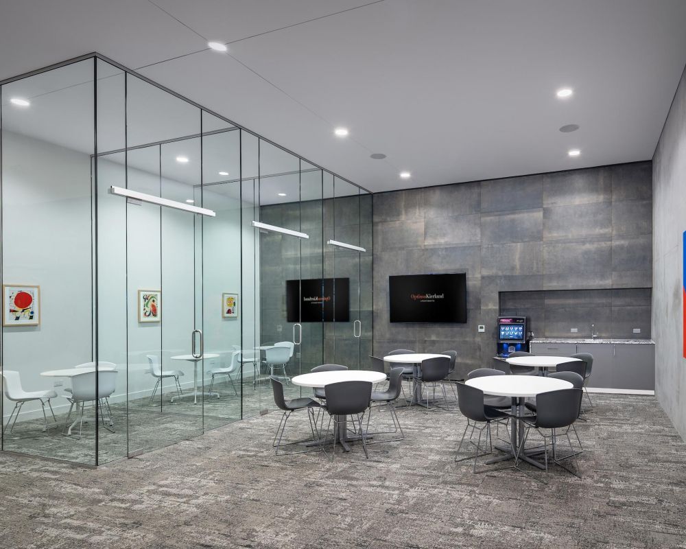 Modern office break room with round tables, gray chairs, glass-walled meeting room, and wall art.