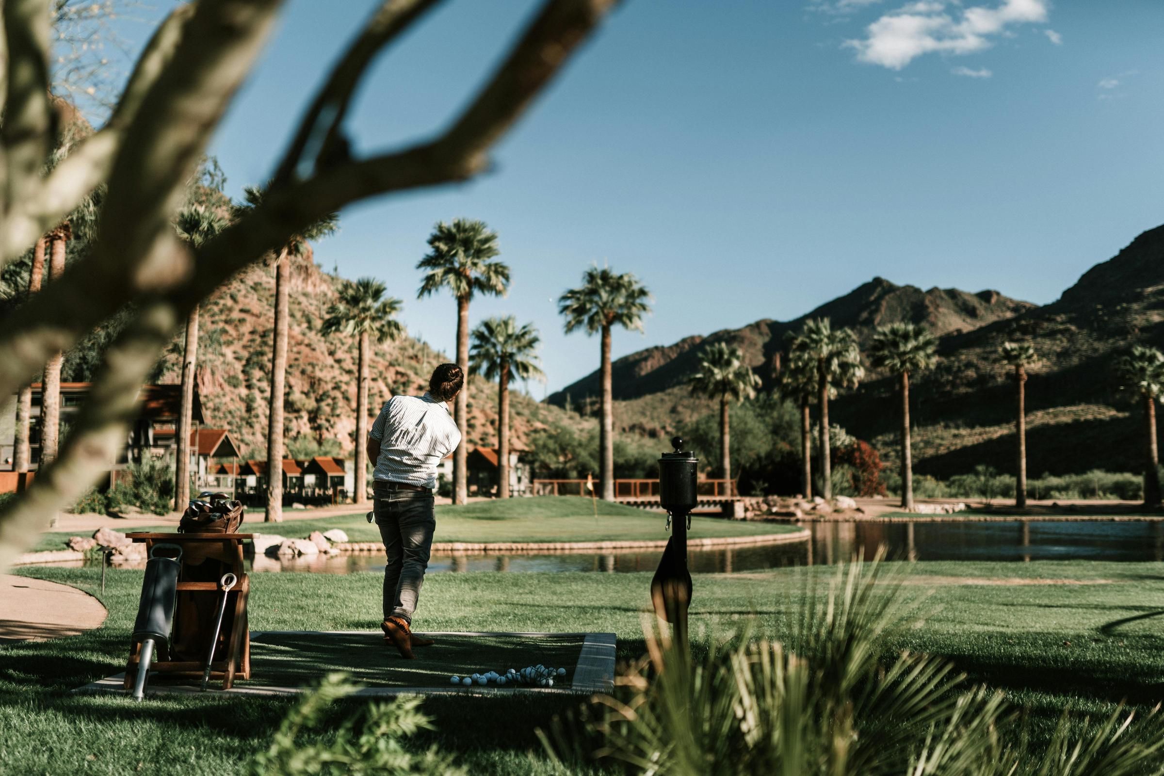 Person golfing in a scenic landscape with palm trees, mountains, and a lake under a clear blue sky near Optima Sonoran Village apartments in Scottsdale, AZ.