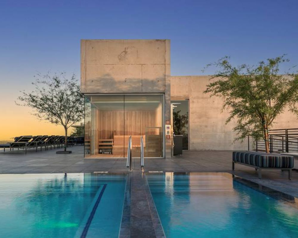 Modern building with large glass windows and a pool in front, surrounded by trees and lounge chairs at sunset.