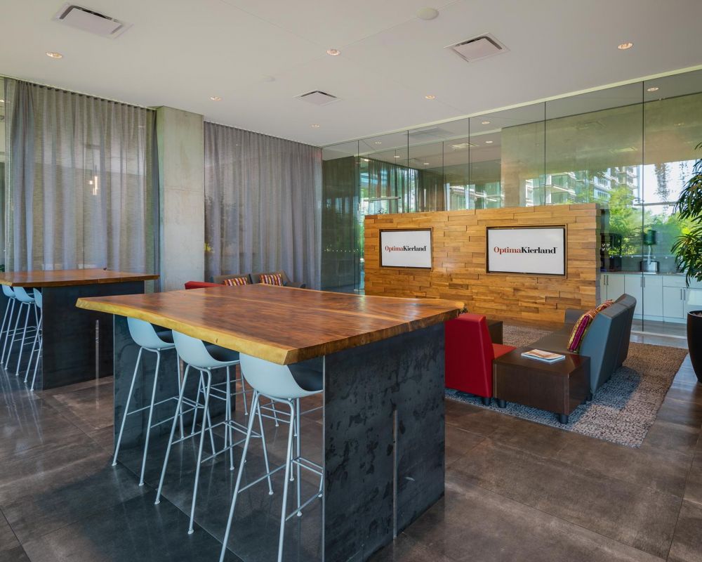 Modern office lounge with tall tables, white chairs, glass walls, and a cozy seating area with TV screens.