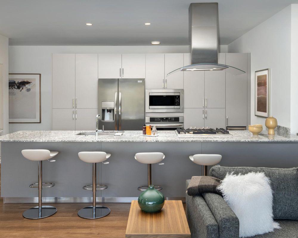 Modern kitchen with gray cabinets, stainless steel appliances, and four white bar stools at a marble island.