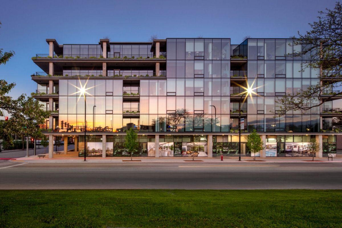 Modern glass architecture reflecting sunset, with trees and street in the foreground, bathed in natural light.